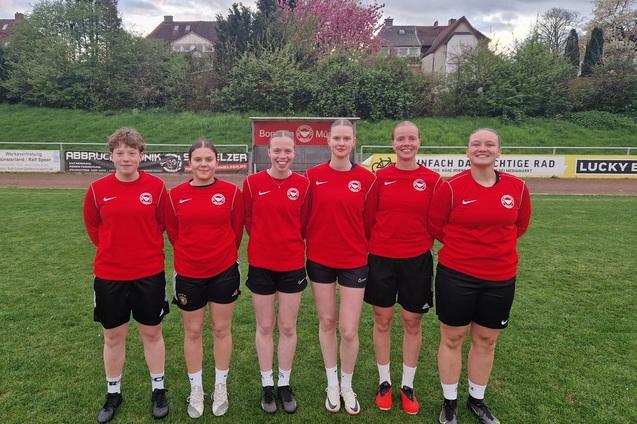 Sechs Fu&szlig;ballspielerinnen in roten Trikots stehen auf einem Sportplatz, im Hintergrund B&auml;ume und eine Werbetafel.