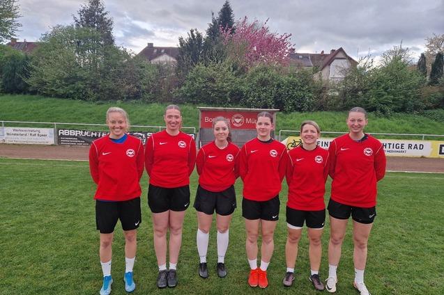 Sechs Fu&szlig;ballerinnen in roten Trikots stehen auf einem gr&uuml;nen Platz, im Hintergrund B&auml;ume und eine Stadionwand.