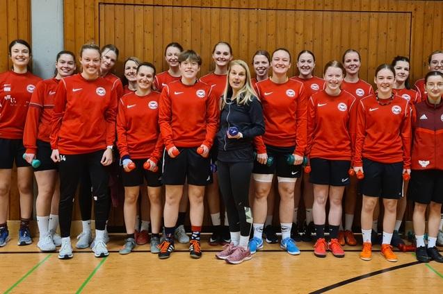 Gruppenfoto von 15 Frauen in roten Trainingsjacken mit Hanteln in der Hand, posing in einer Sporthalle.