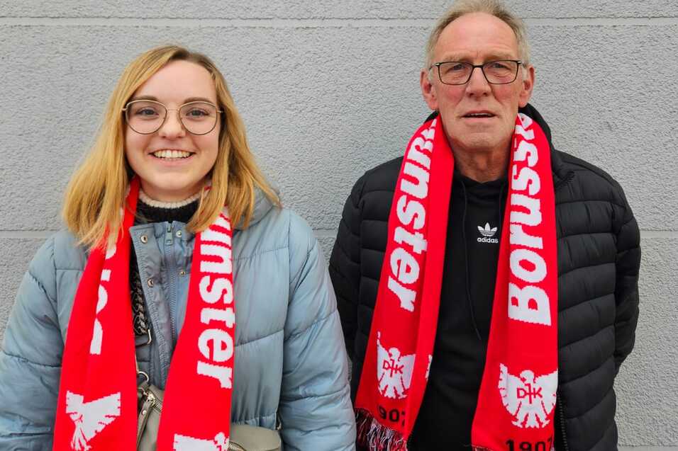 Frau mit blonden Haaren und Brille sowie Mann mit grauen Haaren, beide tragen rote Schal mit "Borussia" auf grauem Hintergrund.