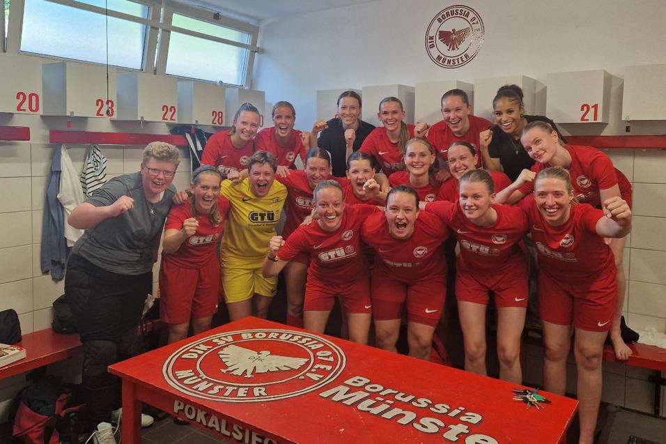 Feiernde Fu&szlig;ballmannschaft in roten Trikots posiert jubelnd in der Umkleidekabine mit Pokalsieger-Schild auf dem Tisch.