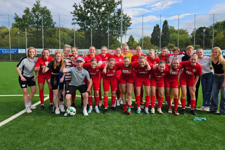 Gruppe von Fu&szlig;ballspielerinnen in roten Trikots posiert lachend auf einem Sportfeld, mit einer Trainerperson in der Mitte.