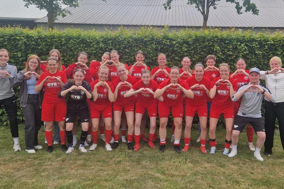 Gruppenzusammenstellung von Frauenfu&szlig;ballspielerinnen in roten Trikots, posierend vor einer gr&uuml;nen Hecke.