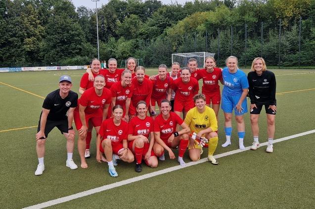 Mannschaftsaufstellung einer Fu&szlig;ballmannschaft in roten Trikots auf einem Kunstrasenplatz, umgeben von B&auml;umen.