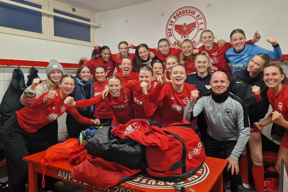 Gruppe von Frauen in Rot, jubelnd in einer Umkleidekabine, mit Trikots und Taschen auf einem Tisch.
