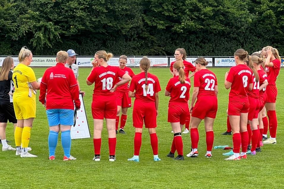 Zehn Frauen im roten Fu&szlig;balltrikot stehen um eine Trainerin, die Strategie auf einem Taktikboard erkl&auml;rt.