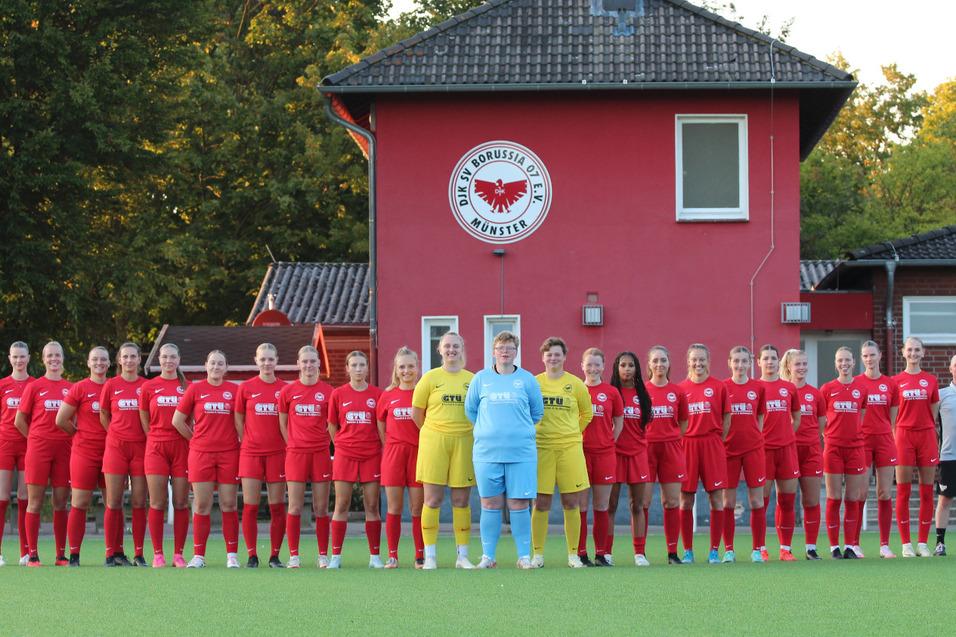 Gruppenfoto einer Frauenfu&szlig;ballmannschaft in roten Trikots vor einem Vereinsgeb&auml;ude mit Wappen.