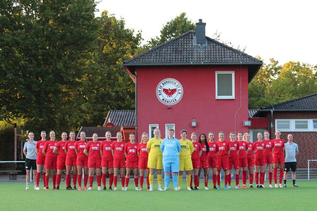 Gruppenfoto einer Frauenfu&szlig;ballmannschaft in roten Trikots vor einem Vereinsgeb&auml;ude mit Wappen.