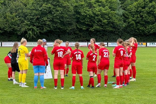 Zehn Frauen im roten Fu&szlig;balltrikot stehen um eine Trainerin, die Strategie auf einem Taktikboard erkl&auml;rt.