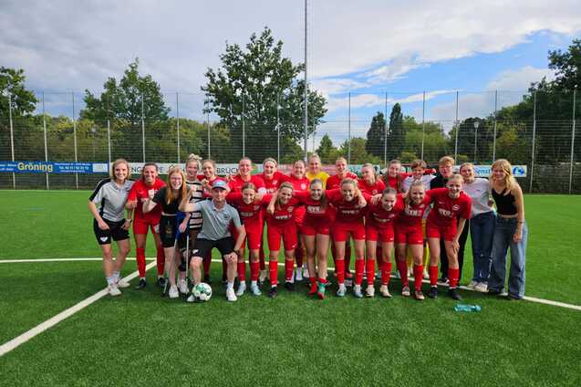 Gruppe von Fu&szlig;ballspielerinnen in roten Trikots posiert lachend auf einem Sportfeld, mit einer Trainerperson in der Mitte.