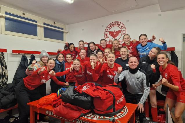 Gruppe von Frauen in Rot, jubelnd in einer Umkleidekabine, mit Trikots und Taschen auf einem Tisch.