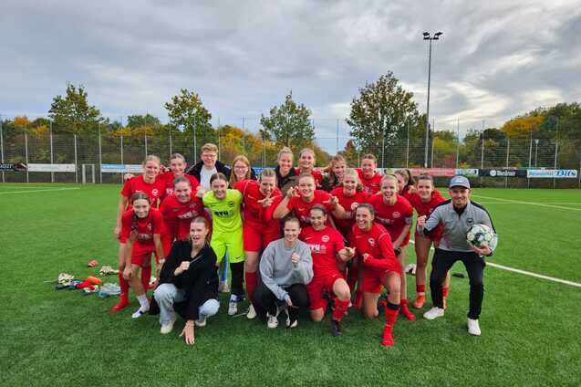 Gruppe von Fu&szlig;ballspielerinnen in roten Trikots posiert fr&ouml;hlich auf einem Kunstrasenplatz nach einem Spiel.