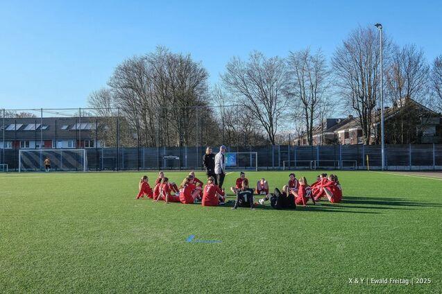 Mannschaft in roten Trikots sitzt auf einem Fu&szlig;ballfeld, umgeben von B&auml;umen und einem Sportgel&auml;nde im Hintergrund.