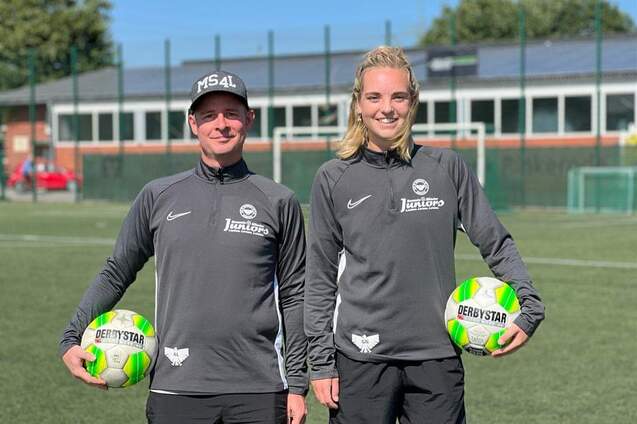 Zwei Trainer in grauen Trainingsanz&uuml;gen halten je einen Fu&szlig;ball auf einem Sportplatz mit gr&uuml;ner Rasenfl&auml;che.