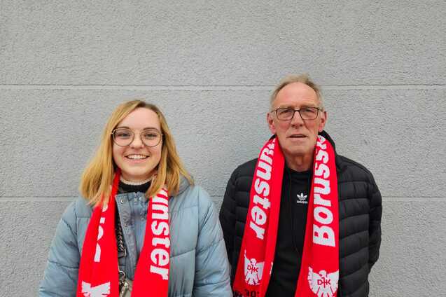 Frau mit blonden Haaren und Brille sowie Mann mit grauen Haaren, beide tragen rote Schal mit "Borussia" auf grauem Hintergrund.