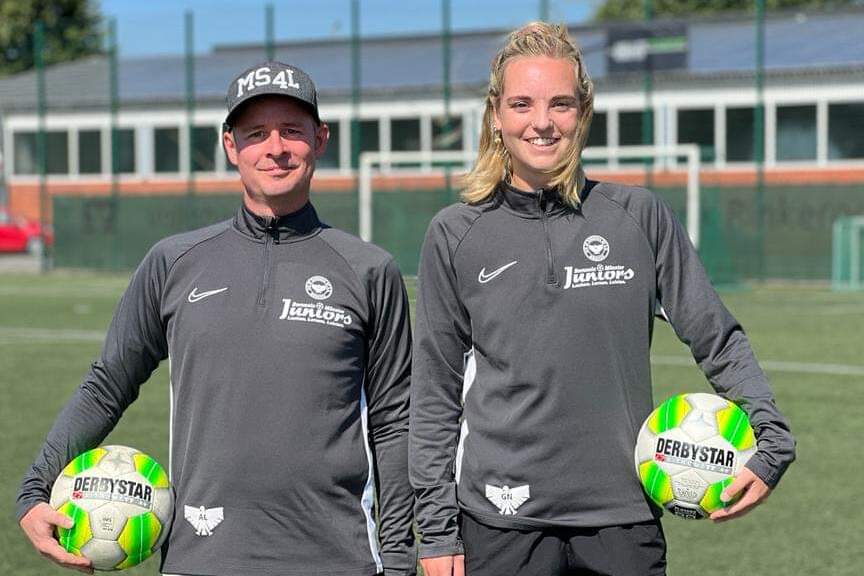 Zwei Trainer in grauen Trainingsanz&uuml;gen halten je einen Fu&szlig;ball auf einem Sportplatz mit gr&uuml;ner Rasenfl&auml;che.