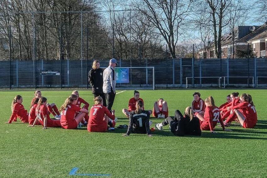 Mannschaft in roten Trikots sitzt auf einem Fu&szlig;ballfeld, umgeben von B&auml;umen und einem Sportgel&auml;nde im Hintergrund.