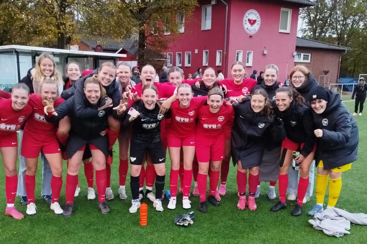 Gruppe von Frauen in rot-schwarzen Fu&szlig;balltrikots posiert fr&ouml;hlich auf einem Rasenplatz vor einem Sportgeb&auml;ude.