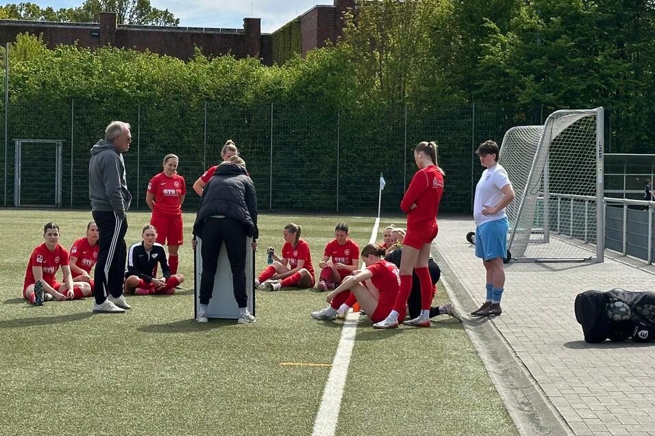 Trainer spricht mit einer Gruppe Fu&szlig;ballspielerinnen in roten Trikots auf einem Kunstrasenplatz.