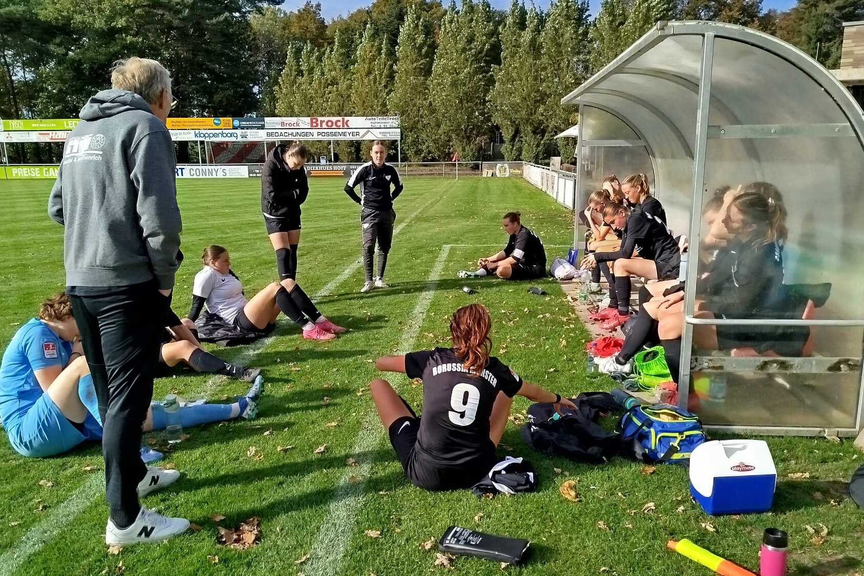 Sitzende und stehende Fu&szlig;ballspielerinnen besprechen sich auf einer Rasenfl&auml;che neben einer Trainerbank.