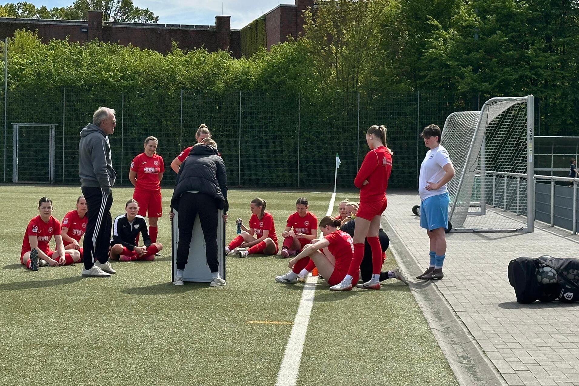Trainer spricht mit einer Gruppe Fu&szlig;ballspielerinnen in roten Trikots auf einem Kunstrasenplatz.