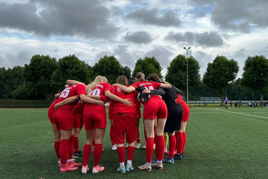 Eine Frauenfu&szlig;ballmannschaft in roten Trikots steht umarmt auf einem Kunstrasenfeld unter wolkigem Himmel.