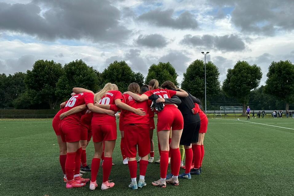 Eine Frauenfu&szlig;ballmannschaft in roten Trikots steht umarmt auf einem Kunstrasenfeld unter wolkigem Himmel.