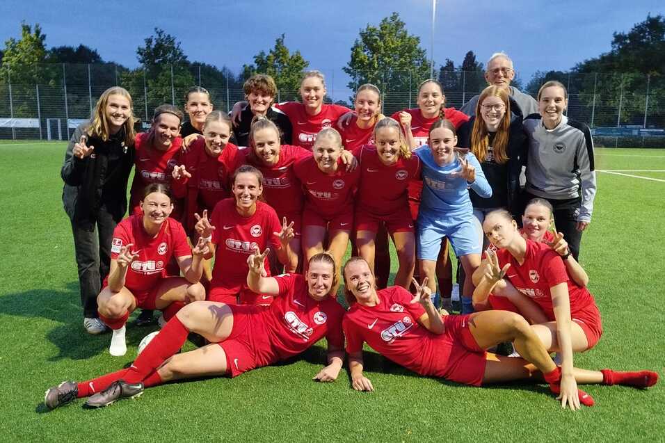Gruppenfoto eines Frauenfu&szlig;ballteams in roten Trikots, feiernd auf einem Kunstrasenplatz unter Flutlicht.