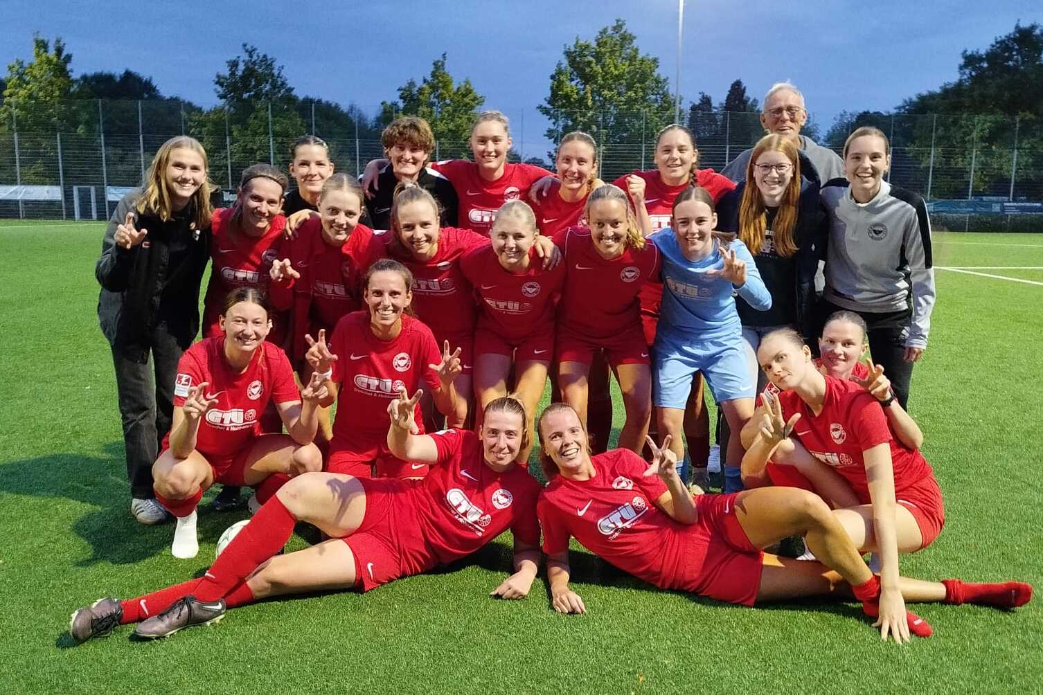Gruppenfoto eines Frauenfu&szlig;ballteams in roten Trikots, feiernd auf einem Kunstrasenplatz unter Flutlicht.