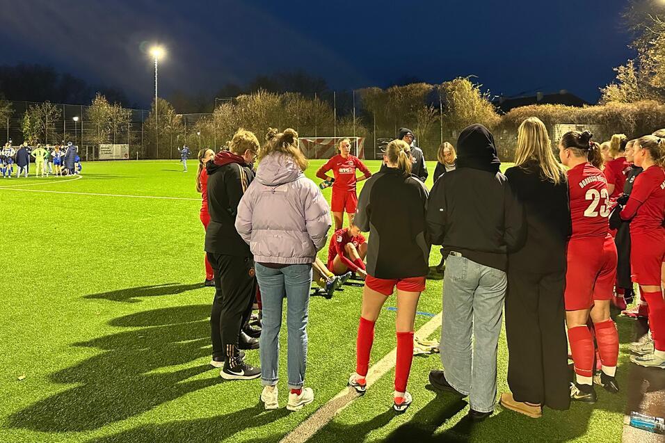 Gruppensitzung von Spielerinnen in roten Trikots mit Trainer auf einem Fu&szlig;ballfeld unter Flutlicht bei D&auml;mmerung.