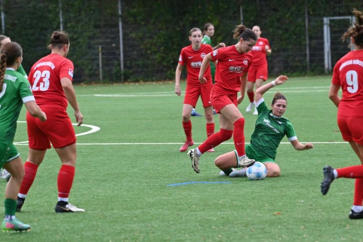 Vier Frauen in roten Trikots spielen Fu&szlig;ball gegen eine Frau in gr&uuml;nem Trikot auf einem Kunstrasenplatz.