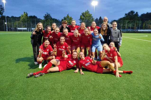 Gruppenfoto eines Frauenfu&szlig;ballteams in roten Trikots, feiernd auf einem Kunstrasenplatz unter Flutlicht.