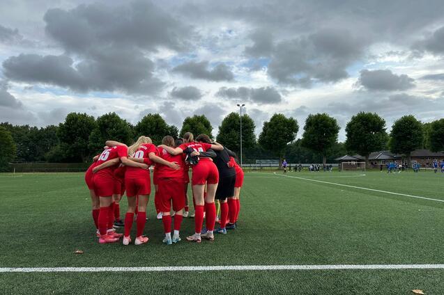 Eine Frauenfu&szlig;ballmannschaft in roten Trikots steht umarmt auf einem Kunstrasenfeld unter wolkigem Himmel.