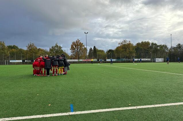 Mannschaft huddelt sich auf einem Kunstrasenplatz vor einem Fu&szlig;ballspiel, im Hintergrund B&auml;ume und grauer Himmel.