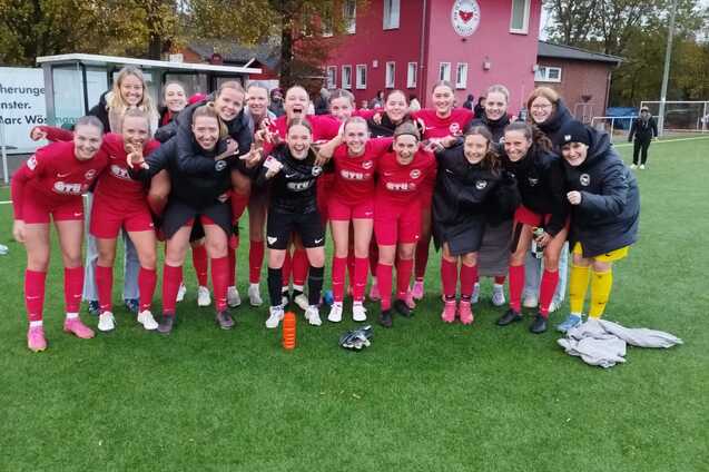 Gruppe von Frauen in rot-schwarzen Fu&szlig;balltrikots posiert fr&ouml;hlich auf einem Rasenplatz vor einem Sportgeb&auml;ude.