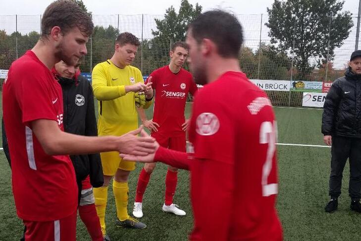 Fu&szlig;ballspieler in roten Trikots sch&uuml;tteln sich zum Spielbeginn die H&auml;nde auf einem Kunstrasenplatz.