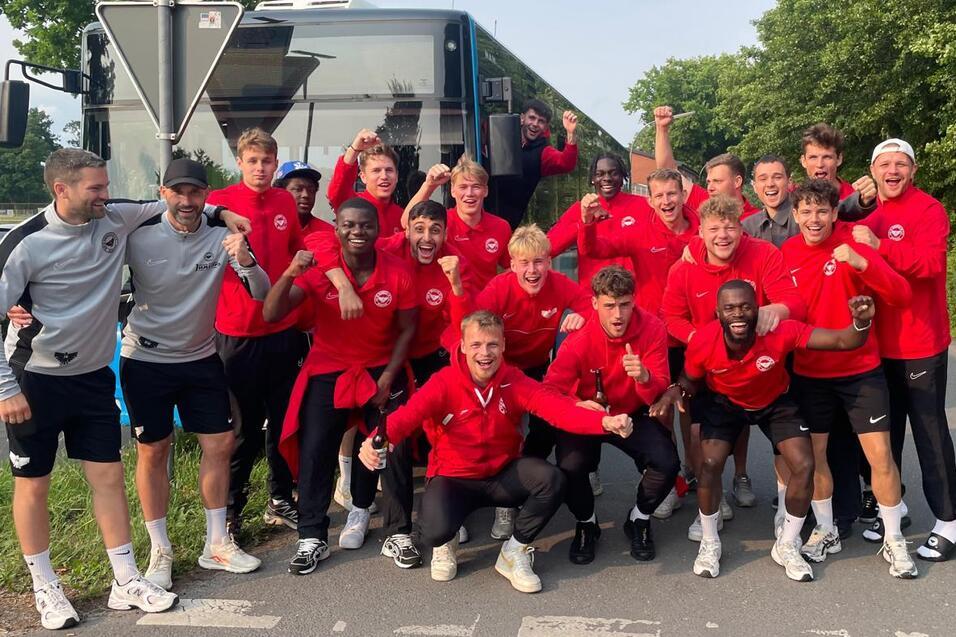 Gruppenselfie von jungen Sportlern in roten Trainingsanz&uuml;gen vor einem Bus an einer Stra&szlig;enecke.