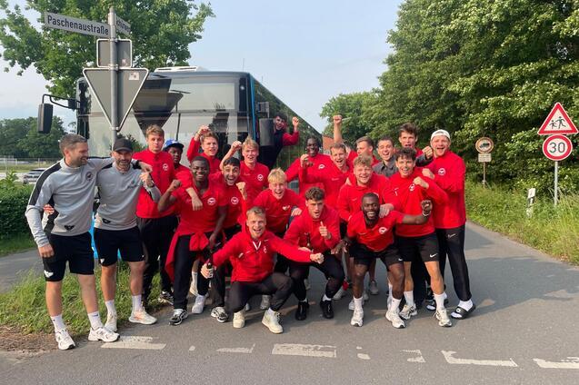 Gruppenselfie von jungen Sportlern in roten Trainingsanz&uuml;gen vor einem Bus an einer Stra&szlig;enecke.