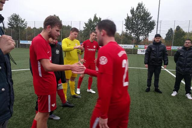 Fu&szlig;ballspieler in roten Trikots sch&uuml;tteln sich zum Spielbeginn die H&auml;nde auf einem Kunstrasenplatz.