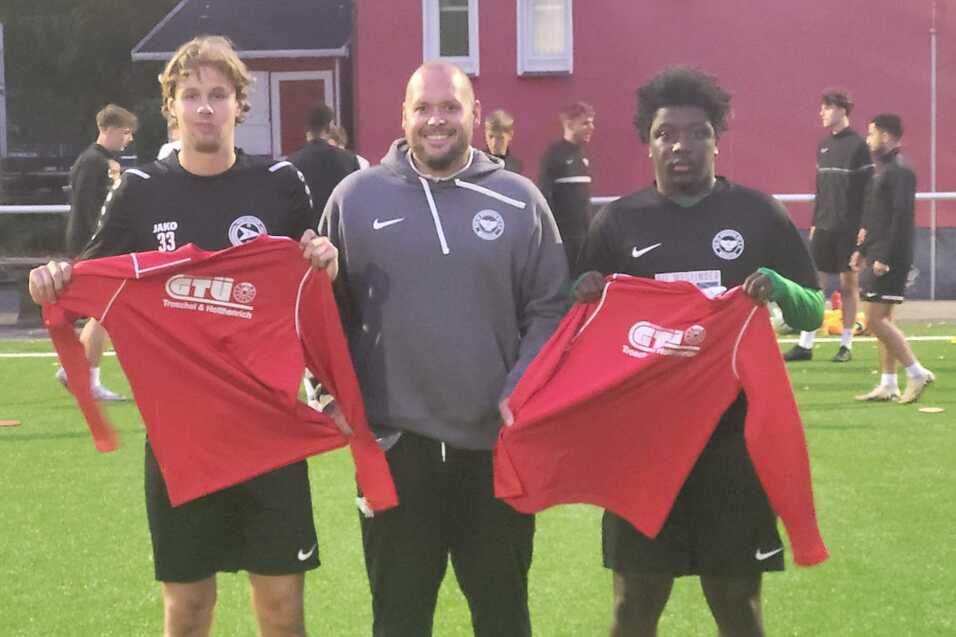 Drei M&auml;nner auf einem Fu&szlig;ballfeld halten rote Trikots; im Hintergrund sind weitere Sportler beim Training zu sehen.