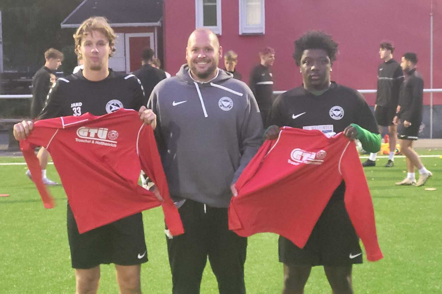 Drei M&auml;nner auf einem Fu&szlig;ballfeld halten rote Trikots; im Hintergrund sind weitere Sportler beim Training zu sehen.