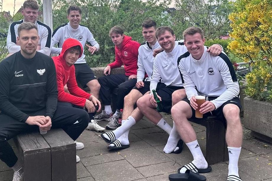 Gruppe von acht M&auml;nnern in Sportbekleidung sitzt entspannt auf einer Terrasse, einige halten Getr&auml;nke in den H&auml;nden.