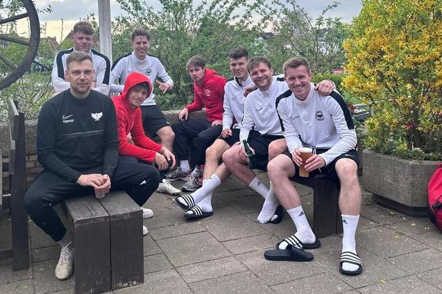 Gruppe von acht M&auml;nnern in Sportbekleidung sitzt entspannt auf einer Terrasse, einige halten Getr&auml;nke in den H&auml;nden.