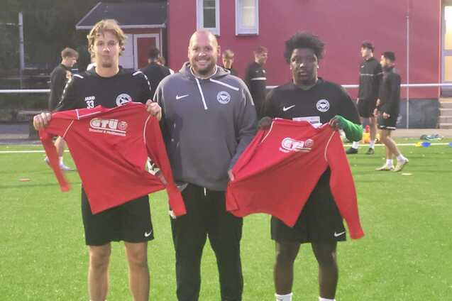 Drei M&auml;nner auf einem Fu&szlig;ballfeld halten rote Trikots; im Hintergrund sind weitere Sportler beim Training zu sehen.