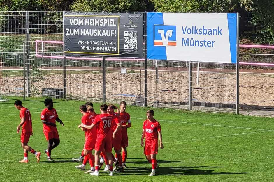 Spieler des Fu&szlig;ballteams in roten Jerseys feiern ein Tor auf einem gr&uuml;nen Sportplatz. Im Hintergrund stehen Tore und ein Werbeschild.
