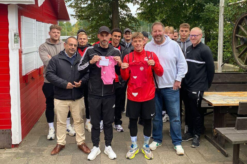 Gruppe von M&auml;nnern und Jugendlichen posieren vor einem roten Geb&auml;ude, ein Junge h&auml;lt ein Trikot in der Hand.
