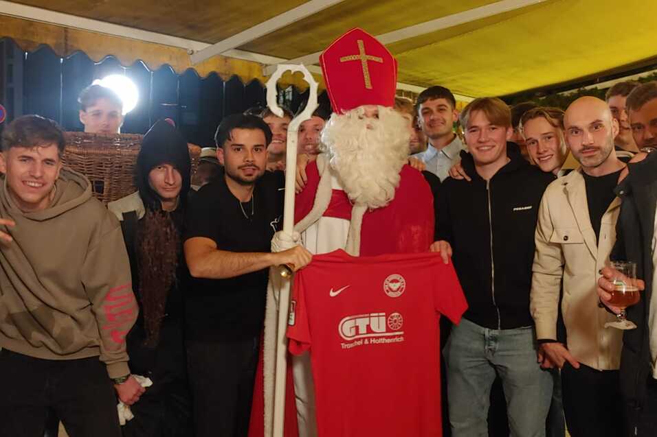 Gruppe junger Menschen posiert mit einem Santa Claus, der ein rotes T-Shirt h&auml;lt, in einer festlich dekorierten Umgebung.