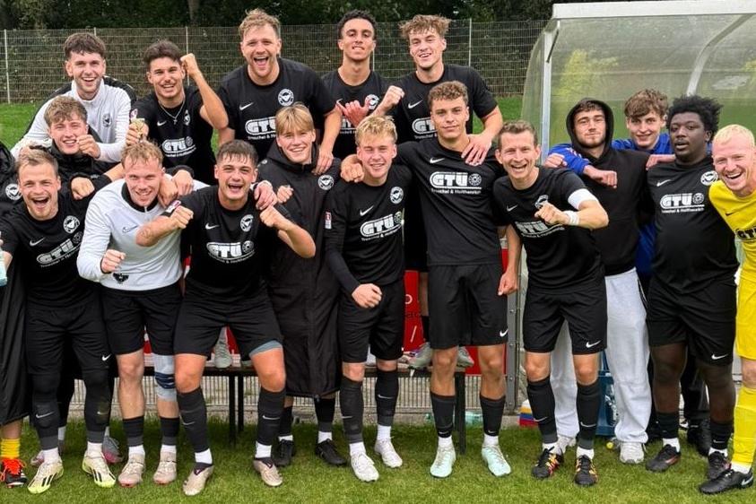 Gruppe von Fu&szlig;ballspielern in schwarzen Trikots steht jubelnd auf dem Spielfeld, einige tragen spezielle Trainingsbekleidung.
