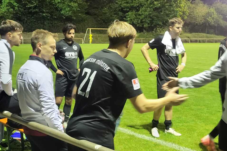 Eine Gruppe von Fu&szlig;ballspielern und Trainern sitzen an der Seitenlinie eines Fu&szlig;ballfeldes bei Nacht.
