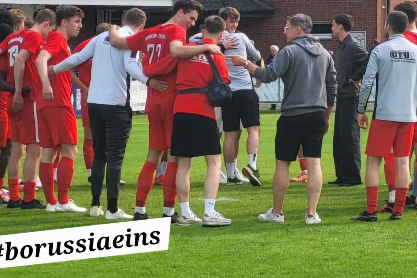 Gruppe von Spielern im roten Trikot versammelt auf einem Sportplatz, im Hintergrund ein Trainer und Banner mit Infos zum Training.